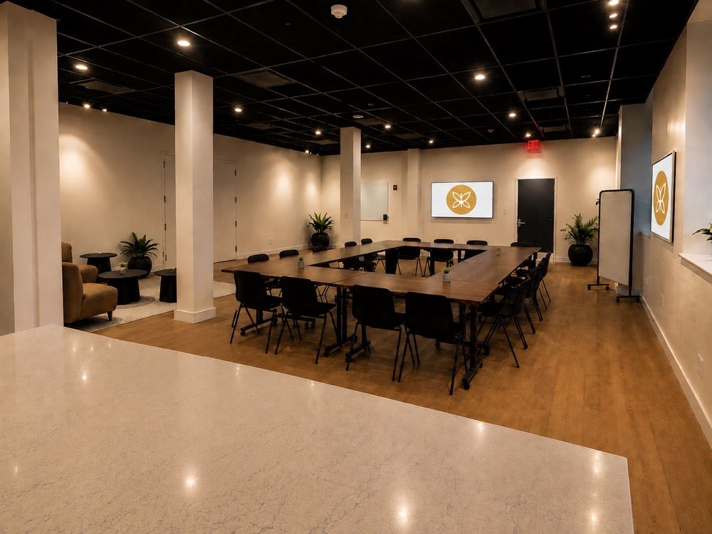 The main room at Monarch Well: tables arranged in a wide open square with seating for about twenty, a lounge corner to one side, and the Monarch Well mark on the screen.