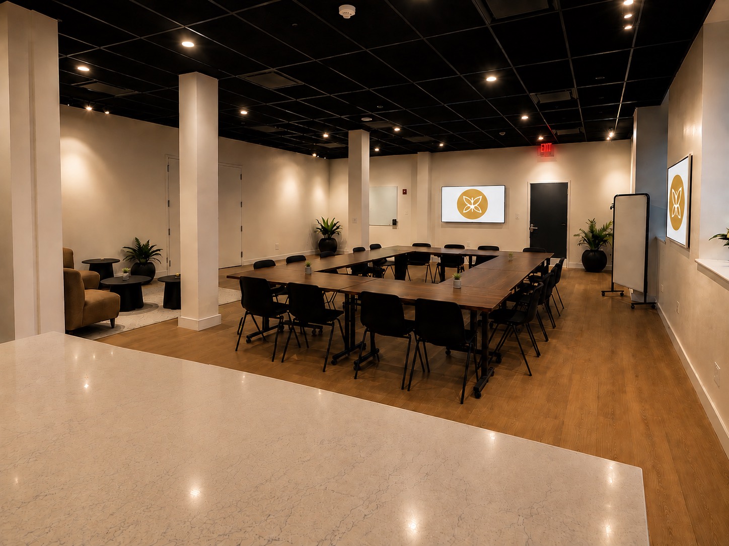 The main room at Monarch Well: tables arranged in a wide open square with seating for about twenty, a lounge corner to one side, and the Monarch Well mark on the screen.