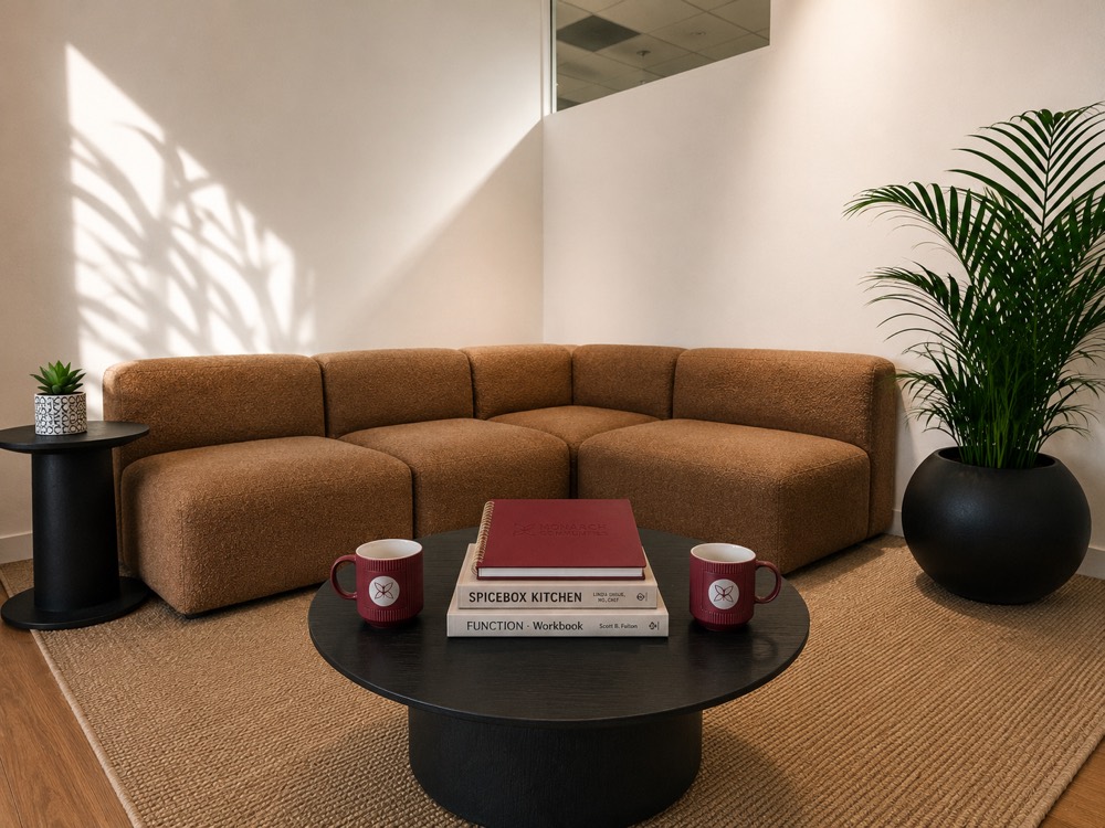 A boucl&eacute; sofa and low table in the Monarch Well lounge, with a stack of books and two Monarch Well mugs set out for a small group.